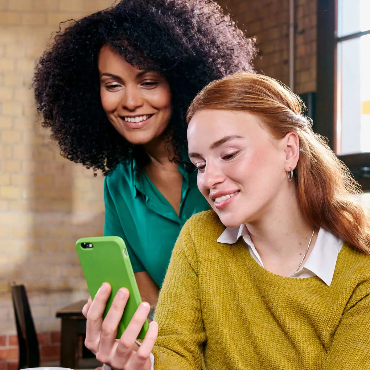 Zwei lächelnde Frauen, eine mit dunkelbraunen lockigen Haaren und eine mit roten Haaren, schauen gemeinsam auf ein Smartphone mit grüner Hülle.